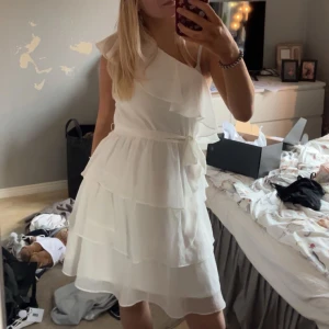 Studentklänning - Superfin vit kortklänning med omlott och volangdetaljer. Klänningen har oneshoulder och ett knytband i midjan som ger en snygg siluett. Perfekt för sommarens alla tillfällen!  Perfekt till student/examen.Den är en 38 tror jag och jag är en 36, syns inte på bilden men den är lite stor över brösten men lätt att antigen sy in eller sätta en nål🩷🩷 