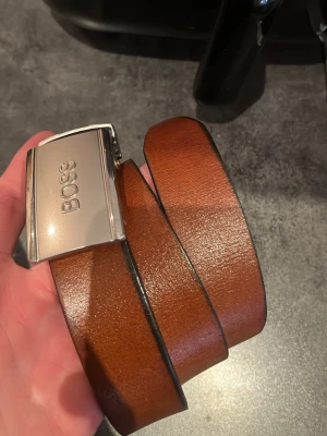 Brunt skärp från BOSS med silverdetalj - Snyggt brunt skärp från BOSS med stilren, blank silverfärgad spänne där BOSS-loggan är ingraverad. Skärpet är gjort i slätt skinn och har svarta kanter för en extra clean look. Perfekt accessoar för att lyfta outfiten.