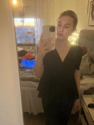 Svart omlottblus med v-ringning - Säljer en svart blus med omlott-design och v-ringad hals. Toppen har korta ärmar och ett lätt utställd midjeparti som ger en snygg siluett. Perfekt att styla med både jeans och kjol.