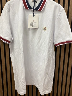 Vit tröja med rödblå krage - Säljer en klassisk vit tröja med korta ärmar. Kragen och ärmsluten har snygga rödblå ränder . Tröjan är gjord i bomull och har en normal passform. Perfekt till en clean och sopad look.