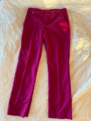 Fuchsia byxor från Ralph Lauren - Snygga fuchsiafärgade byxor från Ralph Lauren med raka ben och klassisk passform. Byxorna har bälteshällor, sidofickor och en liten RL-detalj i metall vid linningen. Tillverkade i ett mjukt bomullsmaterial med lite stretch för extra komfort.
