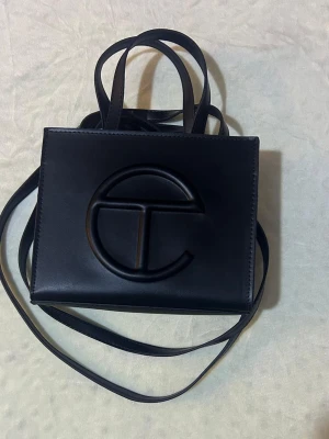 Svart axelväska från Telfar - Säljer en svart axelväska från Telfar med det ikoniska T-logot på framsidan. Väskan har både handtag och lång axelrem, samt ett fack med dragkedja inuti. Tillverkad i slätt syntetmaterial som ger en clean och modern look.