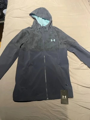 Under Armour grå vindjacka med huva - Under Armour vindjacka i mörkgrå och ljusgrå toner med camo-mönstrad övre del och ljusblå insida på huvan. Jackan har hel dragkedja, två sidofickor och en snygg logga på bröstet. Perfekt för dig som gillar sportig stil och funktion. Fick jackan i julklapp så den är aldrig använd endast testad. Säljer pga att den är för stor