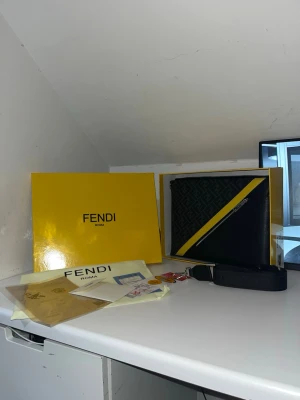 Fendi svart och gul axelväska - Säljer en snygg kuvertväska från Fendi med klassiskt FF-mönster i mörkgrått och svart, samt en diagonal gul detalj och silverfärgad dragkedja. Väskan är tillverkad i skinn och har en avtagbar axelrem med Fendi-logga. Kommer med originalbox, dustbag och äkthetsbevis. Topp kvalitet fraktar inom 1-2 arbetsdagar!!!