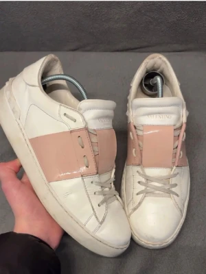 Valentino Garavani vita och rosa sneakers - Säljer ett par Valentino Garavani sneakers i vitt skinn med breda ljusrosa detaljer över vristen. Klassisk rund tå, snörning och platt sula. Märket syns tydligt på plösen och sulan. Snygg och stilren design som sticker ut.