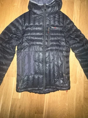 Svart dunjacka från Canada Snow - Svart dunjacka från Canada Snow med huva och flera fickor med dragkedja. Jackan har quiltad design och logga med röd lönnlöv på bröstet. Perfekt för kalla dagar och har en snygg, sportig look. Passar dig som gillar stil och funktion.