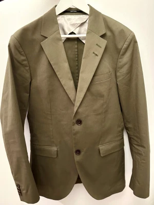 Olivgrön kavaj från Tiger of Sweden, stl 48 - Snygg olivgrön kavaj i ull från Tiger of Sweden med klassisk passform i storlek 48. Kavajen har två knappar framtill, fyra dekorativa knappar vid ärmslut och flera fickor. Perfekt för en stilren look. Extra knappar medföljer.