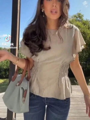 Beige blus med volang och smock - Supersöt beige blus med korta, vida ärmar och smockdetaljer i midjan som ger en snygg siluett. Blusen har rund halsringning och volang nertill för en extra feminin touch. Perfekt att styla med jeans eller kjol för en chill look.