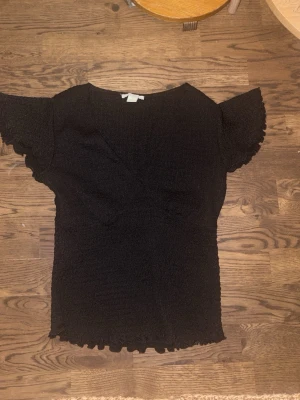 Svart topp med volangärm från Amisu - Säljer en svart topp från Amisu i storlek S. Toppen har korta volangärmar och en smockad, stretchig struktur som sitter snyggt på kroppen. Den har en lätt v-ringad omlottlook och vågig kant nertill för extra detalj.