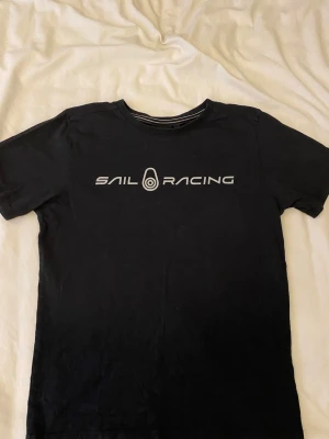 Svart Sail Racing t-shirt i bomull - Svart t-shirt från Sail Racing med vit logga tryckt över bröstet. Klassisk rund halsringning med diskret randig detalj på insidan av kragen. Skön och enkel modell i mjuk bomull, perfekt till jeans eller shorts.