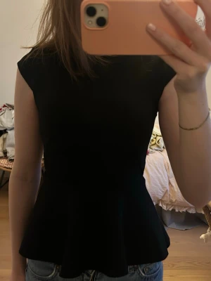 Svart peplum topp  - Snygg svart topp med peplumskärning och capärm. Toppen har en figurnära passform upptill och en utsvängd nederdel som ger en fin siluett. Perfekt att styla med jeans eller kjol för en trendig look.🩷