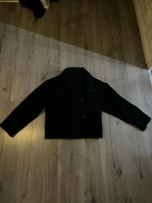 Marinblå kort kappa med knappar - En stilren marinblå kappa med klassisk krage och två knappar framtill. Jackan är rak i modellen och har långa ärmar. Perfekt för dig som gillar en enkel och clean look. Materialet känns mjukt och lite tjockare, vilket gör den nice till kyligare dagar.