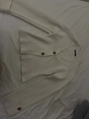 Krämvit kofta från Gina Tricot - Säljer en stilren beige kofta från Gina Tricot i storlek XS. Koftan har v-ringning, långa ärmar och snygga knappar framtill. Perfekt att slänga över en topp för en enkel men chic look.