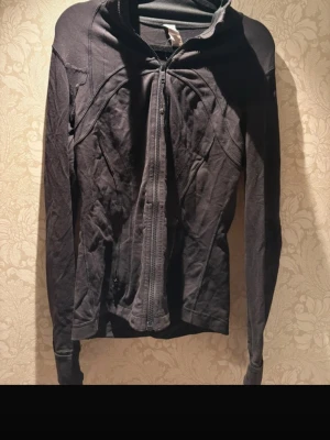 Svart träningsjacka från Lululemon - Säljer en svart, figurnära träningsjacka från Lululemon med dragkedja framtill och snygga sömdetaljer. Jackan har hög krage, långa ärmar med tumhål och diskret logga på ryggen. Perfekt för dig som vill ha en sportig och stilren look.Den sitter som en s men står storlek 8! 