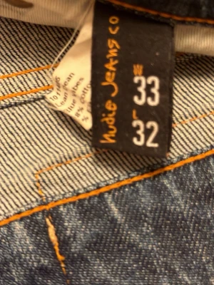Blå jeans från Nudie Jeans, W33 L32 - Säljer ett par klassiska blå jeans från Nudie Jeans med orange kontrastsömmar och den ikoniska vågade brodyren på bakfickorna. Jeansen har rak passform och är tillverkade i slitstarkt bomullsmaterial. Perfekta för dig som gillar tidlös denimstil.