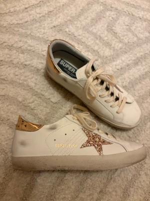 Golden Goose vita sneakers med glitterstjärna - Golden Goose sneakers i vitt skinn med beige snörning, guldmetallic detalj på hälen och en glittrig stjärna i roséguld på sidan. Klassisk låg modell med rund tå och platt sula. Perfekta för dig som vill ha något extra till vardagslooken. Inte mycket använda alls och superfint skick! Pris går absolut att diskuteras. Storlek 38💓