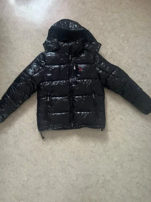 Svart pufferjacka från Polo Ralph Lauren - Säljer en svart, glansig pufferjacka från Polo Ralph Lauren i storlek S. Jackan har huva, flera dragkedjeförsedda fickor och en liten röd logga på bröstet. Perfekt för kalla dagar och riktigt snygg till streetwear-looken.