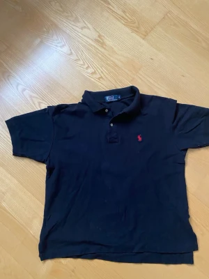 Svart pikétröja från Ralph Lauren XL - Klassisk svart pikétröja från Polo Ralph Lauren i storlek XL. Men den sitter som en M/L. Tröjan har korta ärmar, krage med två knappar och den ikoniska röda broderade loggan på bröstet. Tillverkad i mjuk bomull som känns skön mot huden.