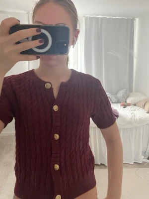 Vinröd kabelstickad kofta med guldknappar -  vinröd kabelstickad kofta med korta ärmar och snygga guldknappar framtill. Koftan har rund halsringning