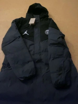 Svart lång pufferjacka PSG x Jordan - Säljer en svart och mörkgrå lång pufferjacka från Jordan x PSG. Jackan har huva, två långa dragkedjor framtill och loggor på bröstet. Perfekt för kalla dagar och har en riktigt clean look med snygga detaljer. 