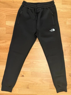 Svarta mjukisbyxor från The North Face - Svarta mjukisbyxor från The North Face med dragsko i midjan och dragkedjeförsedda fickor. Byxorna har en slim passform och är tillverkade i mjukt material, perfekt för chill eller träning. Loggan syns tydligt på ena benet.