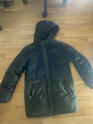 Mörkgrön och svart pufferjacka med huva - Säljer en lång pufferjacka från NN07 i mörkgrönt och svart. Jackan har stor huva, två stora fickor med dragkedja och tryckknappar framtill. Perfekt för kalla dagar och har en clean, stilren look. Märkespatch på ärmen för extra detalj.