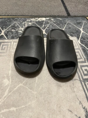 Svarta slides i syntetmaterial - Säljer ett par svarta slides med öppen tå och chunky sula. De har en minimalistisk design och är gjorda i mjukt syntetmaterial som är skönt att gå i. Perfekta för chill dagar eller när du vill ha något enkelt att slänga på fötterna.