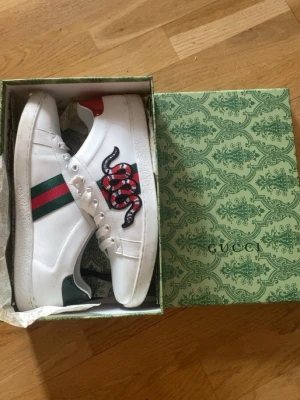 Gucci Ace sneakers med orm - Gucci Ace sneakers i vitt skinn med klassiska gröna och röda ränder på sidan och en broderad orm i rött, svart och vitt. Skorna har vita snören, röd glittrig detalj på hälen och platt sula. Kommer med originalkartong och dustbag.