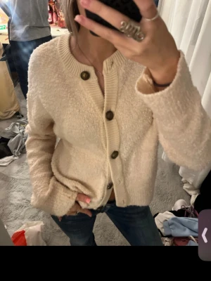 Beige kofta med knappar - Mysig beige kofta med grovstickad känsla och stora bruna knappar framtill. Koftan har lång ärm, ribbad kant vid hals och knappslå samt en loose passform. Perfekt för lager på lager under höst och vinter.