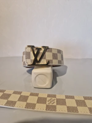 Rutig Louis Vuitton bälte - Säljer ett snyggt bälte från Louis Vuitton med klassiskt rutigt mönster i beige och brunt. Bältet har en stor LV-logga i guld som spänne och är tillverkat i ett tåligt material. Perfekt accessoar för att lyfta din outfit.