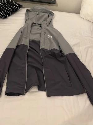 Grå vindjacka  - Säljer en snygg tvåfärgad vindjacka från Under Armour i ljus- och mörkgrått. Jackan har huva, dragkedja framtill, bröstficka med dragkedja och logga på bröstet. Perfekt för blåsiga dagar och sportiga outfits. OBS lite trasigt vid dragkedjan
