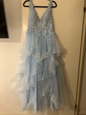 Ljusblå långklänning med spetsdetaljer - Magisk ljusblå långklänning med v-ringning och breda axelband. Överdelen är täckt av blommig spets och kjolen har flera lager volangig tyll som ger en riktigt sagolik känsla. Dragkedja i ryggen för enkel påklädning. Perfekt till bal!! Jag har endast använt den 1 gång, väldigt bra skick! Köpte den online från USA för ett år sedan, då betalade jag ca 3700kr. Hör av dig om du har fler frågor eller funderingar 🥰