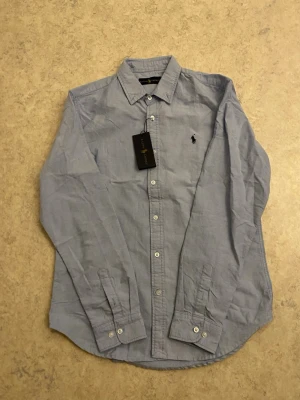 Ljusblå skjorta från Ralph Lauren M - Klassisk ljusblå skjorta från Ralph Lauren i bomull. Skjortan har lång ärm, knappar framtill och den ikoniska broderade loggan på bröstet. Perfekt till jeans eller chinos för en clean och stilren look. Priset kan diskuteras 😊