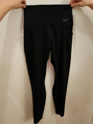 Nya svarta leggings från Nike - Säljer ett par oanvända svarta leggings från Nike med hög midja och sidofickor. Modellen heter nike universa. De är tillverkade i stretchigt syntetmaterial som sitter tight och bekvämt. Perfekta för träning eller chill dagar. Diskret Nike-logga framtill vid midjan.