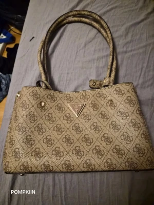 Guess Meridian II shoulder satchel  - Säljer en beige handväska från Guess med klassiskt logomönster och två långa handtag. Väskan har flera fack, inklusive ett med dragkedja i mitten, och stängs med magnetknappar. Triangelformad Guess-logga i metall framtill.