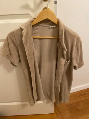 Beige frottéskjorta med kort ärm - Säljer en beige skjorta i mjuk frotté med korta ärmar och klassisk krage. Skjortan har en bröstficka och knäppning framtill. Perfekt för en chill och avslappnad stil.