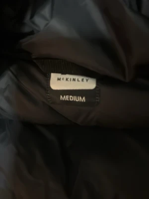 Svart pufferjacka från McKinley - Säljer en svart pufferjacka från McKinley i storlek medium. Jackan har huva, dragkedja framtill och är quiltad med breda kanaler. Perfekt för kalla dagar när du vill hålla dig varm och ändå se stilren ut.