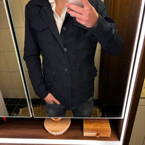 Mörkblå parkajacka från Brandit - Säljer en mörkblå parkajacka från Brandit i storlek M. Jackan har flera praktiska fickor framtill, stängs med både dragkedja och knappar och har en klassisk krage. Materialet känns slitstarkt och passar perfekt till vardagslooken.