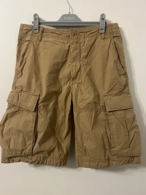 Beiga cargoshorts med stora fickor - Säljer ett par beiga cargoshorts från Cubus med stora sidofickor och klassisk knappstängning. Shortsen har en loose passform och är tillverkade i bomull, perfekta för varma dagar och chill stil.