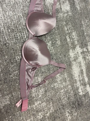 Ljusrosa satin-bh från Juicy Couture - Snygg bh från Juicy Couture i glansigt satinmaterial med ljusrosa utsida och rosa insida. Axelbanden har broderad logga och justerbar längd. Klassisk form med vadderade kupor och hakknäppning bak. Push up. Storlek 34c som motsvarar 75c Lite nopprig. Köpt i usa för 300kr 