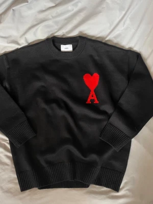 Svart stickad tröja från Ami  - Svart stickad tröja från Ami med rund hals och ribbade muddar. På bröstet finns ett stort rött hjärta med bokstaven A under, vilket ger en unik och trendig look. Perfekt till höst och vinter för dig som gillar stilrena detaljer.