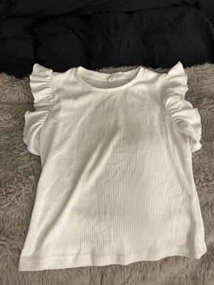 Vit ribbad topp med volangärmar - Säljer en vit ribbad topp med rund hals och stora volangdetaljer på ärmarna. Toppen har en enkel men ändå trendig look och är gjord i ett mjukt, stretchigt material som känns skönt mot huden. Perfekt för dig som gillar en söt och stilren stil.