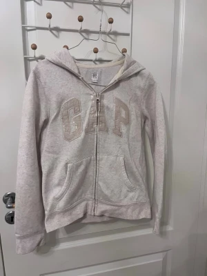 GAP Hoodie - Ljusbeige GAP zip-hoodie i mjukt och bekvämt material. Klassisk modell med dragkedja, huva och fickor framtill. GAP-logga i tyg med diskreta paljettdetaljer som ger en fin detalj utan att bli för mycket. Perfekt som vardagströja eller för mysiga outfits.  