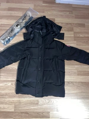 Svart dunjacka från Canada Goose - Säljer en svart dunjacka från Canada Goose med huva och avtagbar pälskant. Jackan har klassisk patch på ärmen, dragkedja och knappar framtill samt flera fickor. Släpper lite dun ibland men perfekt för kalla dagar och riktigt snygg streetstil. Nästan oanvänd.