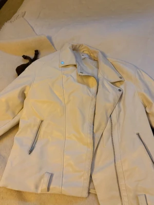 Vit skinnjacka med dragkedjor - Snygg beige skinnjacka från Gina Tricot med coola silverfärgade dragkedjor och detaljer vid kragen. Jackan har en klassisk bikerstil med långärmade ärmar och två fickor med dragkedja framtill. Perfekt för dig som gillar stilrena och trendiga plagg.