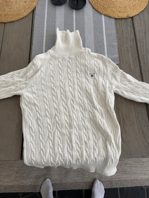 Vit kabelstickad polotröja från Gant M - Säljer en vit kabelstickad polotröja från Gant med hög krage och diskret logga på bröstet. Tröjan har långa ärmar och ribbade muddar vid hals, ärmslut och nederkant. Perfekt för kyliga dagar och riktigt snygg till jeans eller kjol. Storlek M