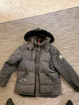 Svart Moose Knuckles dunjacka - Säljer en svart dunjacka från Moose Knuckles med pösig passform och stor huva med fuskpäls. Jackan har flera dragkedjor, tryckknappar och den ikoniska loggan på ärmen. Perfekt för kalla dagar och riktigt snygg till streetwear-stilen.