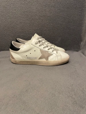 Golden Goose  - Golden Goose sneakers i vitt skinn med grå mockadetaljer och ikonisk stjärna på sidan, i storlek 41. Skorna har används några gånger och har tecken på användning. Nypris 5800. OG är box och boxen medföljer vid köp. Perfekt för varma sommar dagar men också tidig höst och sen vår. Perfekt för dig som vill ha en unik stilren look till vardagen.                                                                Skriv vid frågor, priset är inte hugget i sten🤝