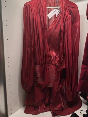 Vinröd omlottklänning  - Säljer en vinröd omlottklänning från Ginatricot med v-ringad hals, långa ärmar och volangdetaljer i nederkant. Klänningen har ett glansigt material och ett brett midjeband . Perfekt för dig som vill sticka ut med en elegant look. Aldrig använd !!!