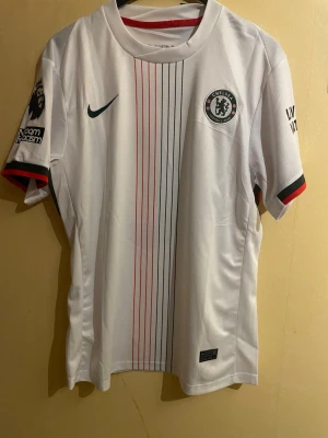 Chelsea bortatröja 2023/24 – Palmer 10 - en sprillans ny Chelsea bortatröja i storlek M. Tröjan är helt oanvänd med tags kvar och i perfekt skick. Stilren vit design med vertikala detaljer, Chelsea-emblem, Premier League-patch samt ”No Room For Racism”-märke. Tryck på ryggen: PALMER 10. Tillverkad i Nike Dri-Fit-material som andas och är bekvämt både till match och vardag. Ett måste för Chelsea-supportern eller samlaren.